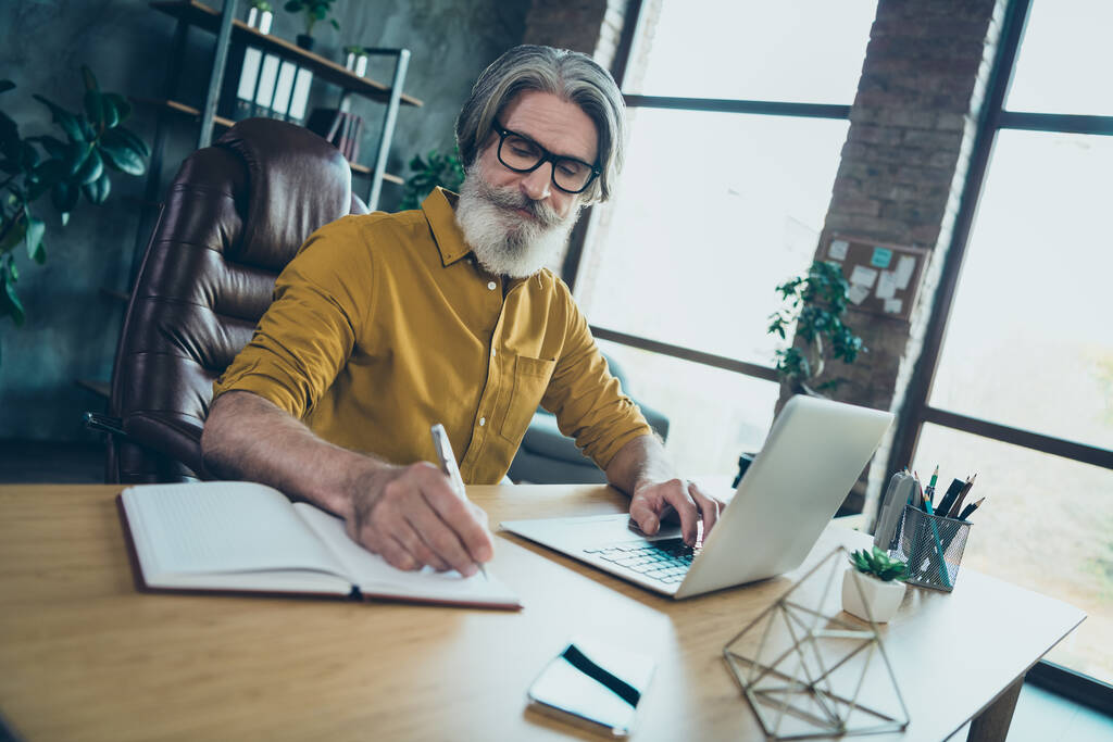 Photo of busy senior man wear yellow shirt glasses typing modern device writing book indoors workshop workplace. - Photo, Image