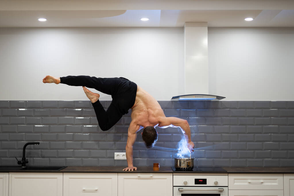 Man keeps balance on one hand and cooks food in the kitchen. Healthy lifestyle, yoga and wellness concept - Photo, Image