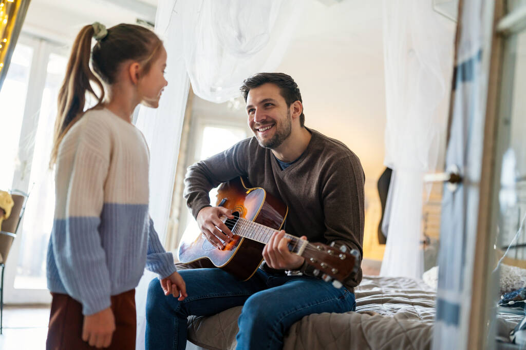 Gelukkig meisje en haar knappe vader spelen gitaar en glimlachen terwijl ze thuis op de bank zitten - Foto, afbeelding