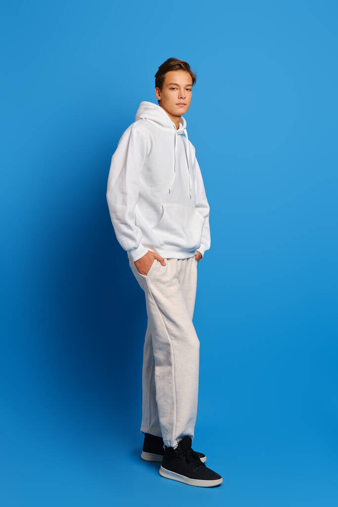 Giovane uomo che indossa felpa bianca e pantaloni della tuta in piedi sopra lo sfondo blu studio con le mani in tasca - Foto, immagini