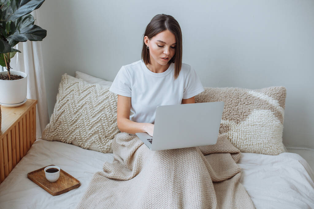Uma jovem morena em uma camiseta branca se senta na cama, coberta com um cobertor e olha para a tela do laptop. No peitoril da janela ao lado das plantas da casa em vasos. Em cima da cama há uma bandeja de madeira com uma caneca branca de café - Foto, Imagem