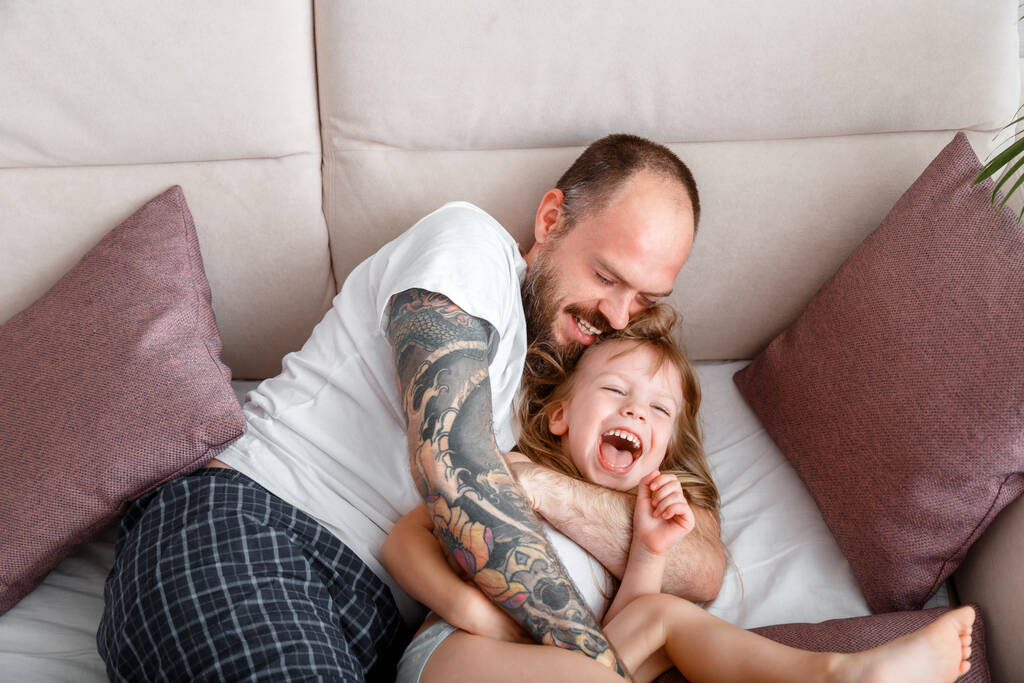 Father play with child kid girl spending time together. Bald bearded Caucasian man in tattoos tickles his tender little daughter. Girl laughs play with dad in pajamas in bed morning routine - Photo, Image