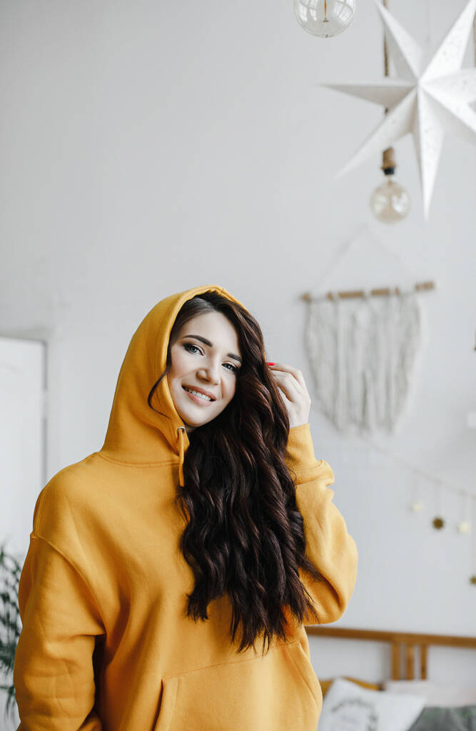 linda chica morena en una sudadera con capucha amarilla con un secador de pelo posa, ríe por un anuncio de salón de belleza - Foto, imagen