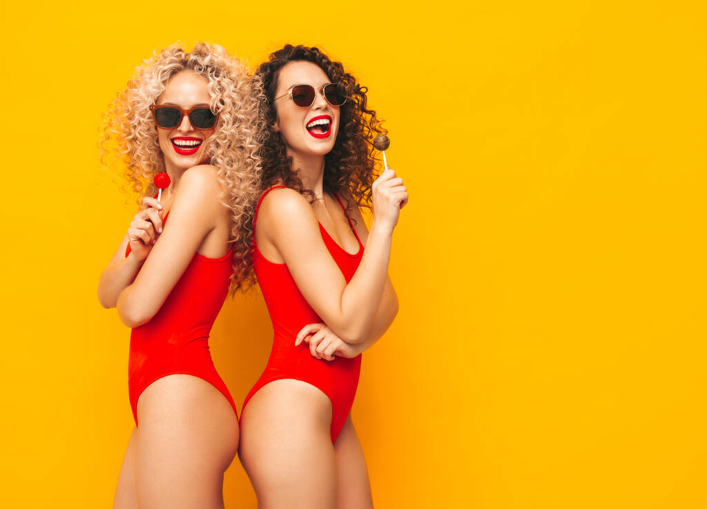 Két gyönyörű mosolygós hipster nők piros fürdőruha fürdőruhában.Trendi modellek fürtök frizura stúdióban. Forró nő pózol közel sárga fal napszemüvegben.Tartása nyalóka. Fehér alapon elszigetelve - Fotó, kép