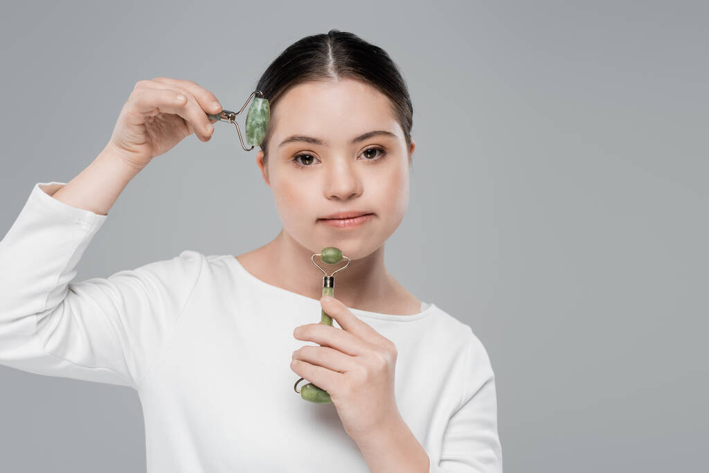 Young woman with down syndrome using jade rollers isolated on grey  - Photo, Image