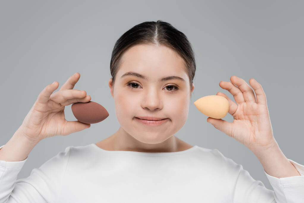 Young woman with down syndrome holding beauty blenders isolated on grey  - Photo, Image