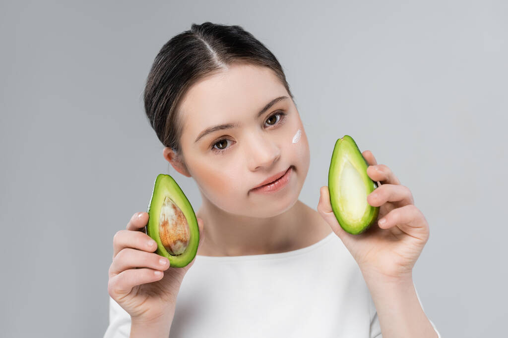 Vrouw met dons syndroom en crème op gezicht houden rijpe avocado geïsoleerd op grijs  - Foto, afbeelding