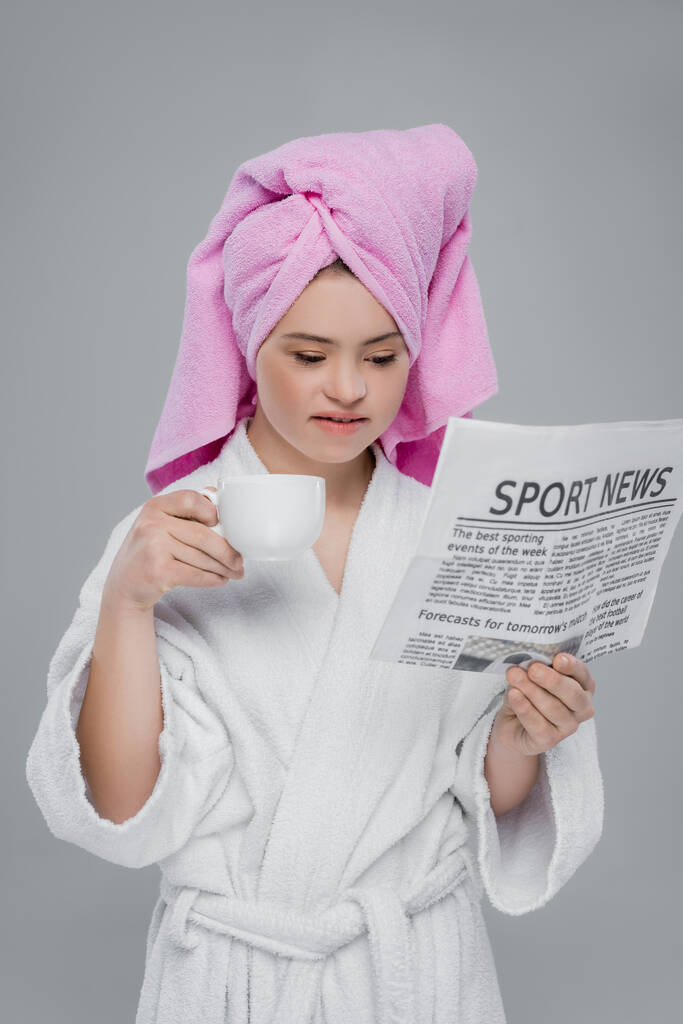 Frau mit Down-Syndrom in Handtuch und Bademantel liest Nachrichten und hält Tasse isoliert auf grau  - Foto, Bild
