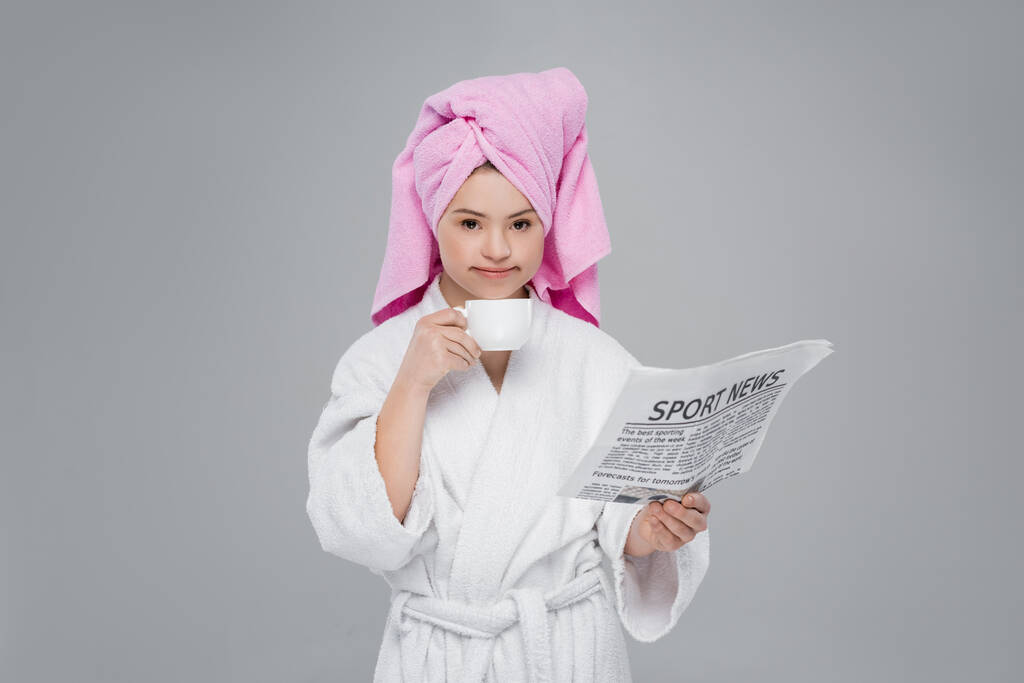 Nainen, jolla on untuvaoireyhtymä kylpytakissa tilalla cup ja sanomalehti eristetty harmaa  - Valokuva, kuva