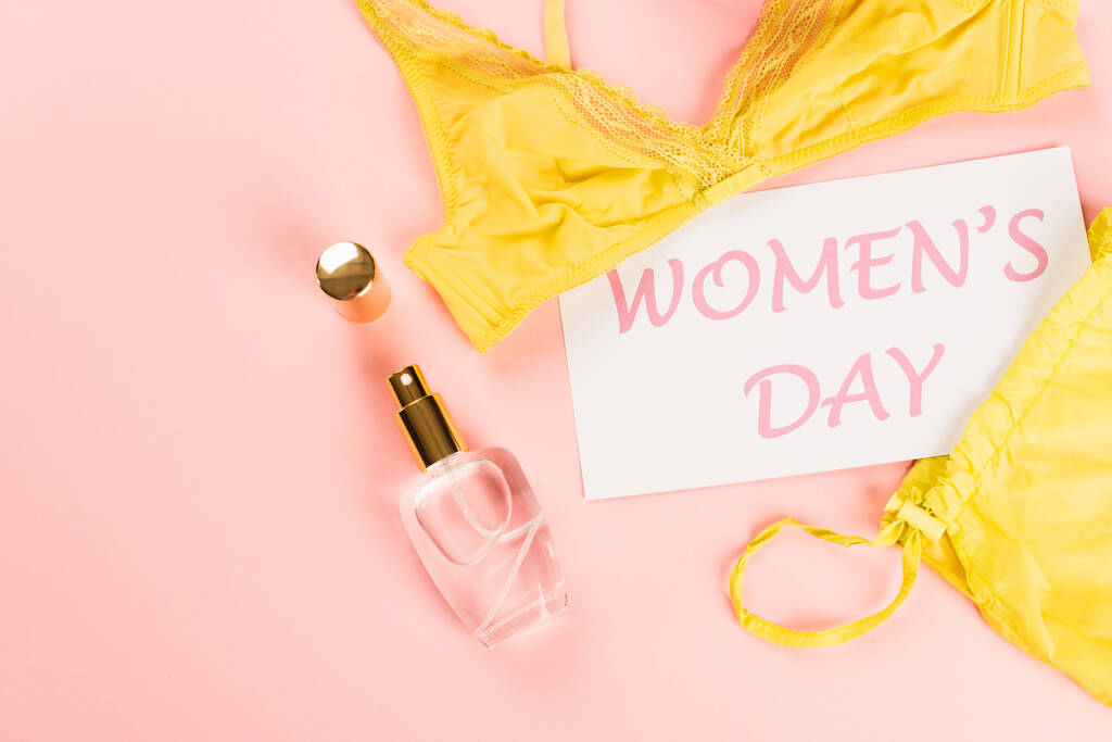 Вид сверху на открытку с женскими дневными нарядами рядом с парфюмом и бюстгальтером на розовом фоне  - Фото, изображение