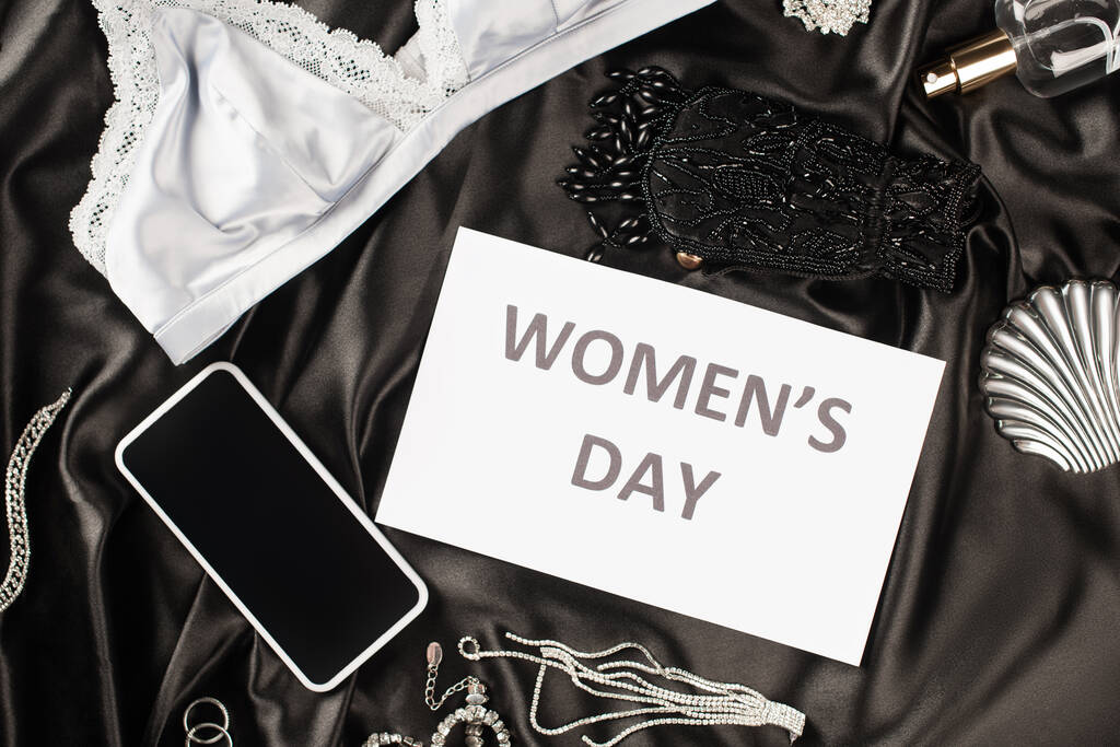 Top view of card with womens day lettering near bra, smartphone and perfume on black satin background  - Photo, Image