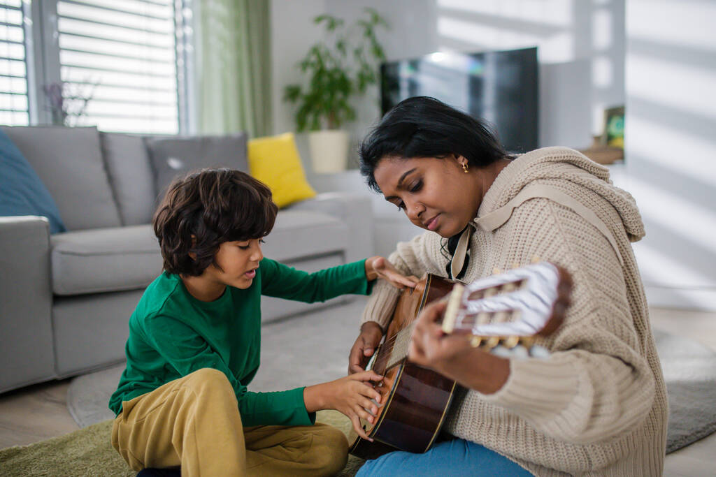 Маленький багаторасовий хлопчик вчиться грати на гітарі з матір'ю вдома
. - Фото, зображення