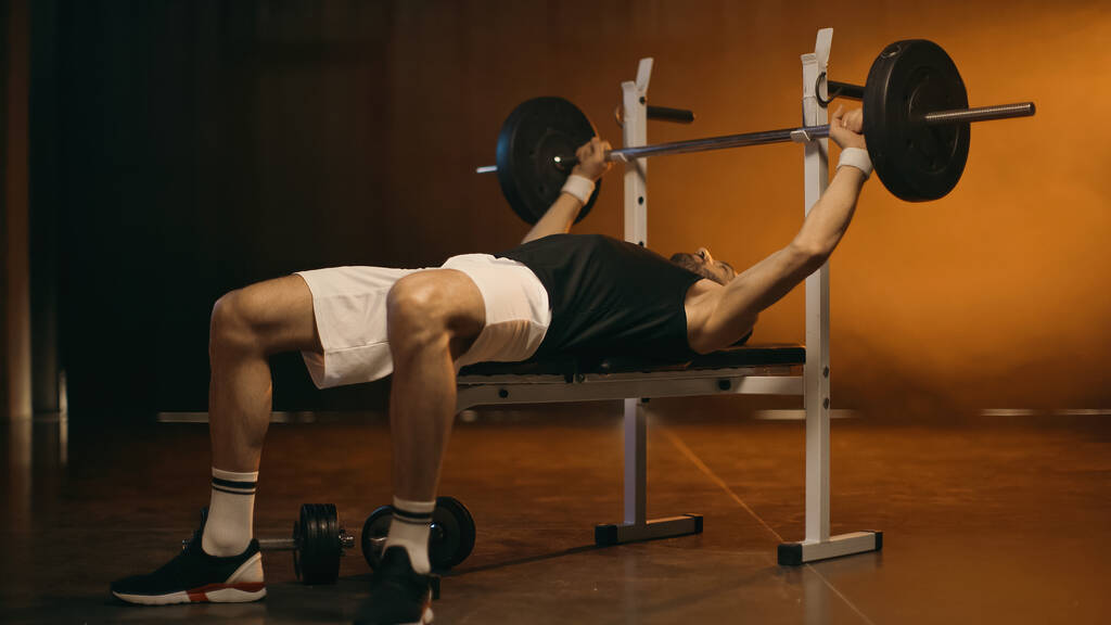 Deportista haciendo ejercicio con barra en banco plano sobre fondo oscuro - Foto, Imagen