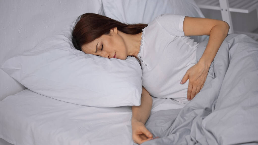 brunette vrouw gevoel pijn in de maag terwijl liggend op bed - Foto, afbeelding
