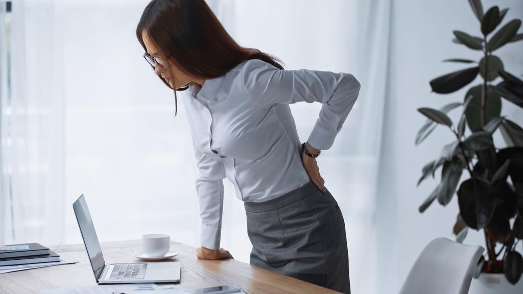 brünette Frau berührt schmerzenden Rücken im Stehen am Schreibtisch - Foto, Bild