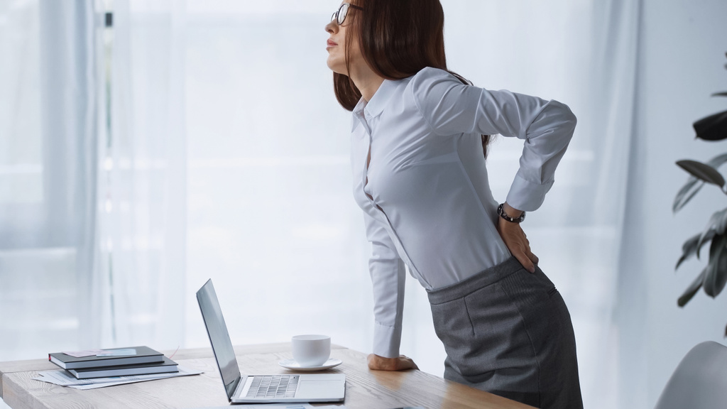brunetka žena stojící za pracovním stolem a dotýkat bolestivé bedra - Fotografie, Obrázek