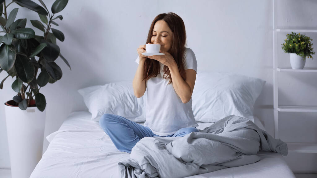 zadowolona kobieta w piżamie siedzi na łóżku i pachnie aromatyczną kawą  - Zdjęcie, obraz