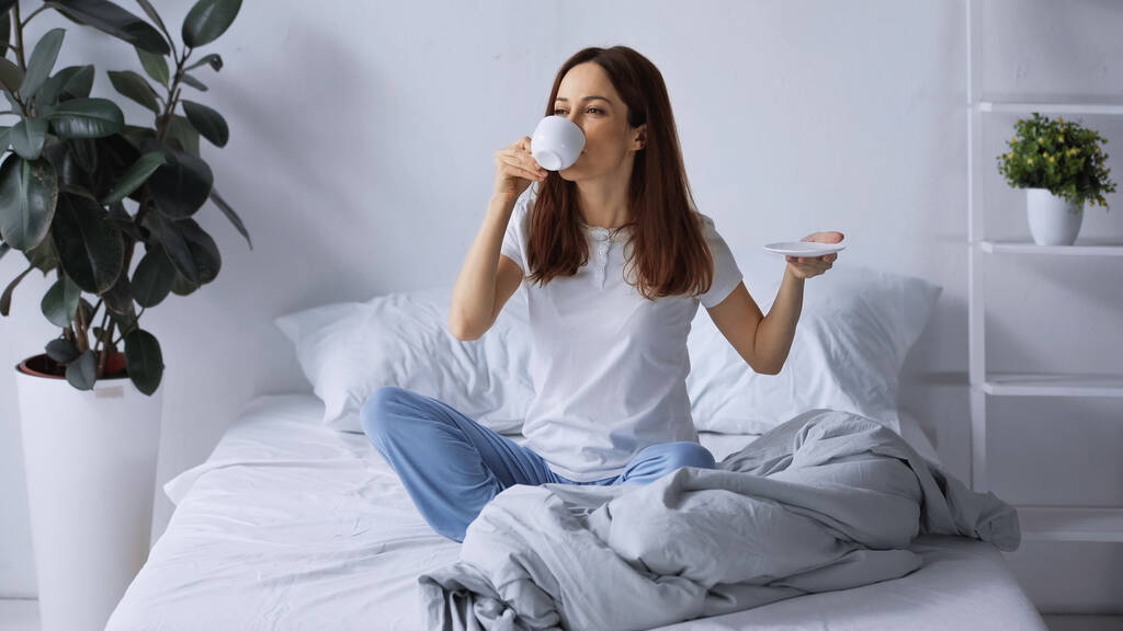 barna nő pizsamában reggeli kávét iszik, miközben az ágyon ül  - Fotó, kép