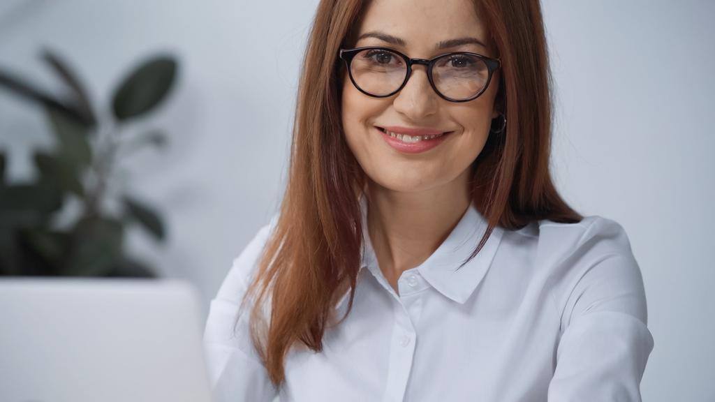 femme d'affaires joyeuse dans les lunettes souriant à la caméra dans le bureau - Photo, image