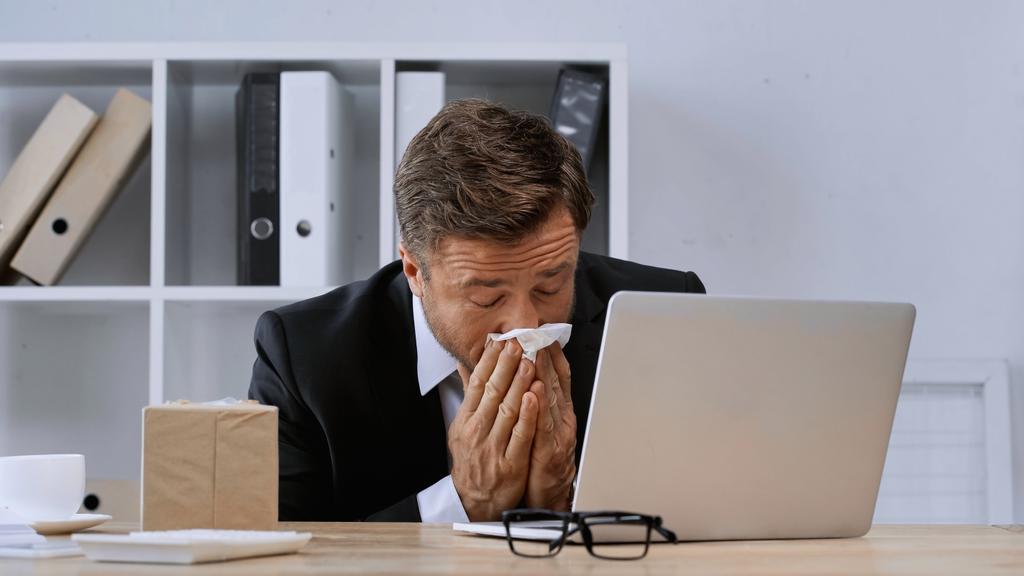 больной бизнесмен с закрытыми глазами чихает в бумажную салфетку возле ноутбука - Фото, изображение