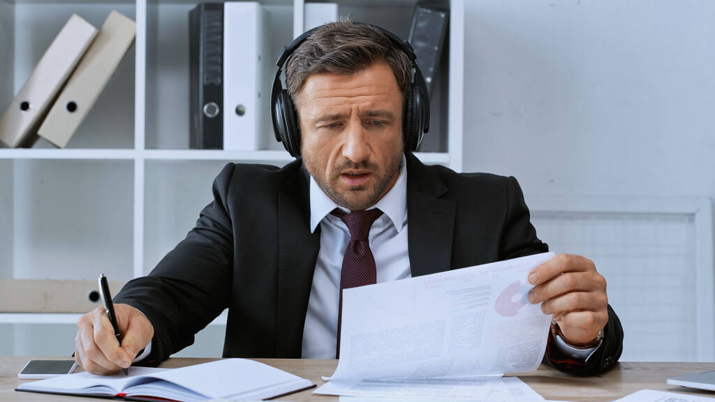 hombre de negocios en auriculares escribiendo en cuaderno mientras trabaja con infografías en la oficina - Foto, Imagen