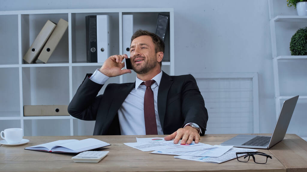 homme d'affaires souriant parlant sur smartphone près d'un ordinateur portable, papiers et tasse de café sur le bureau - Photo, image
