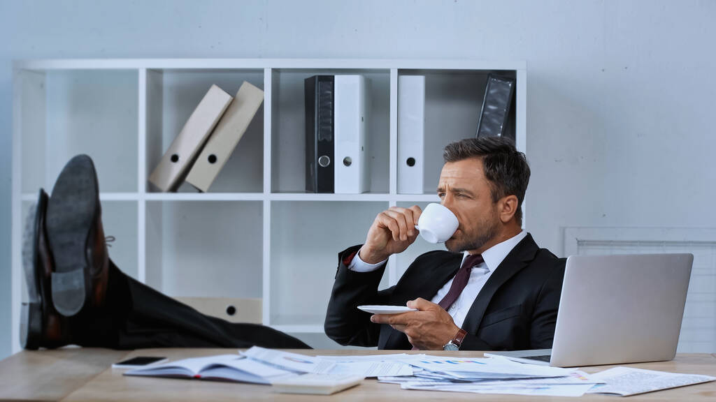 zakenman zit met benen op het bureau tijdens koffiepauze in kantoor - Foto, afbeelding