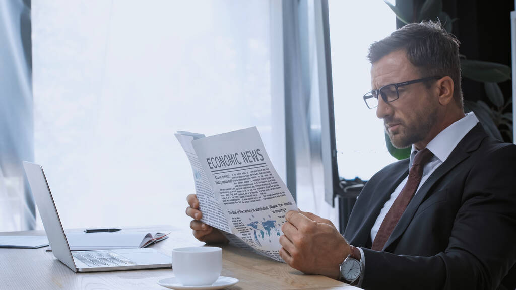 liikemies silmälaseissa lukemassa talousuutisia kahvikupin ja kannettavan tietokoneen lähellä - Valokuva, kuva