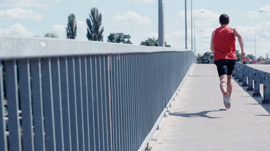 achteraanzicht van een jonge sportman die in de zomer over een stadsbrug rijdt - Foto, afbeelding