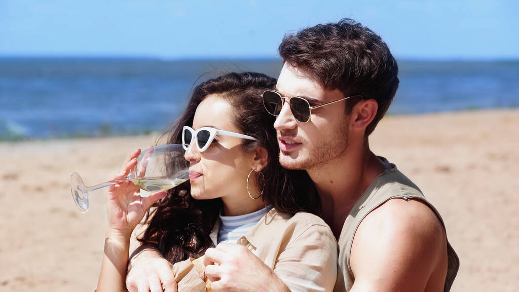 Νεαρή γυναίκα με γυαλιά ηλίου πίνει κρασί ενώ ο φίλος της κρατάει μπαγκέτα στην παραλία  - Φωτογραφία, εικόνα