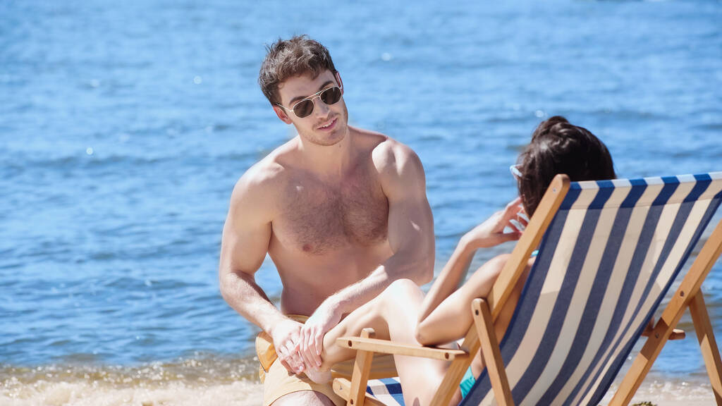 Mosolygó férfi napszemüvegben megérinti a barátnője lábát a fedélzeten szék a strandon  - Fotó, kép