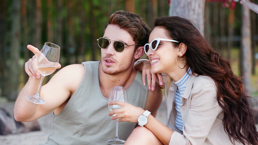 Joven sosteniendo copa de vino y señalando con el dedo a su novia en gafas de sol en la playa  - Foto, imagen