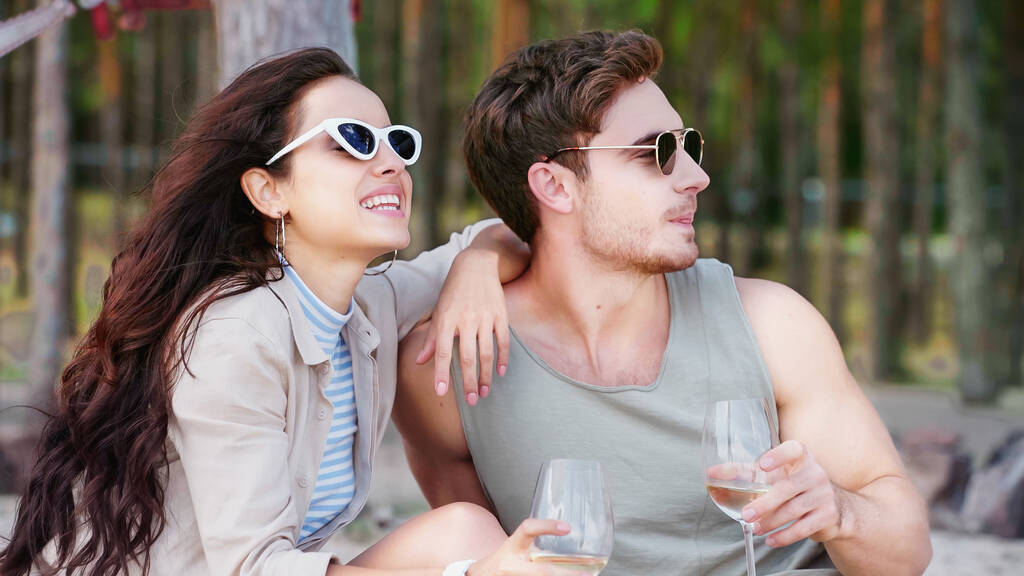 Femme gaie dans des lunettes de soleil tenant un verre de vin près du petit ami sur la plage  - Photo, image