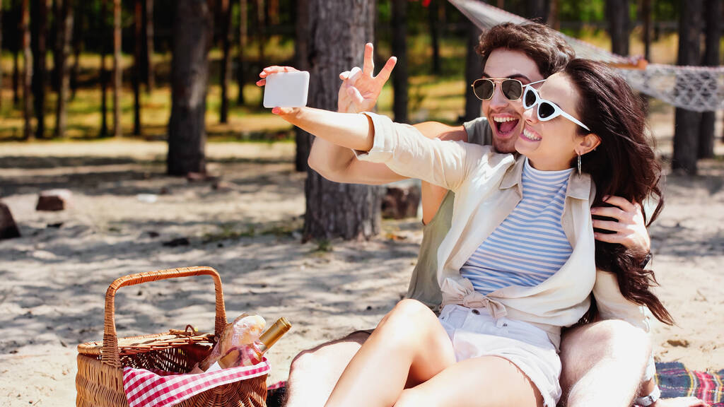 Pareja sonriente tomando selfie en el teléfono inteligente cerca de la cesta de picnic en la playa  - Foto, Imagen