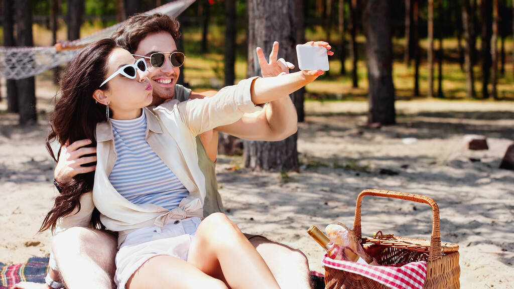 Hymyilevä mies osoittaa rauhan merkki kun selfie tyttöystävä lähellä piknik kori rannalla  - Valokuva, kuva