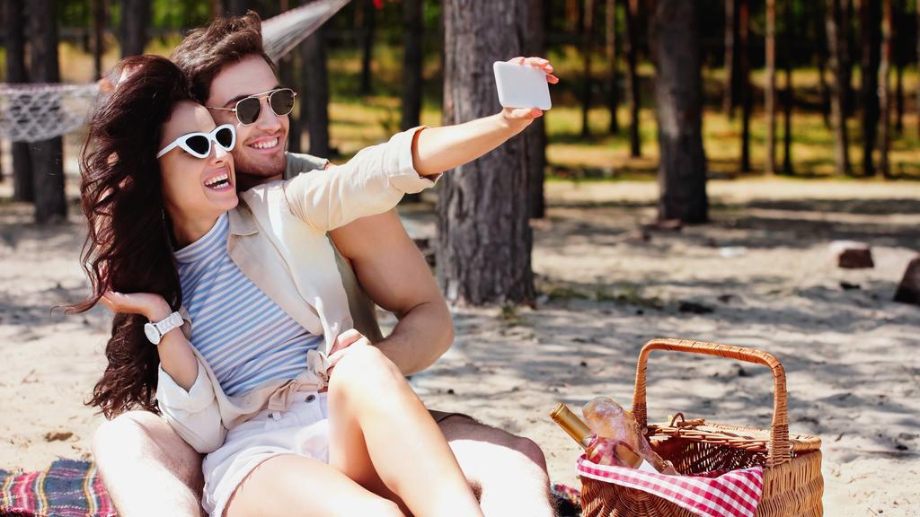 Onnellinen pari aurinkolaseissa ottaen selfie älypuhelimella lähellä koria rannalla  - Valokuva, kuva