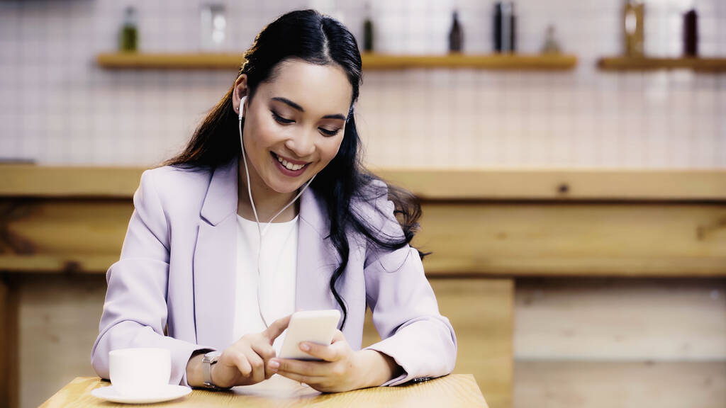 задоволена азіатська жінка слухає музику в навушниках і тримає смартфон біля чашки кави в кафе
 - Фото, зображення