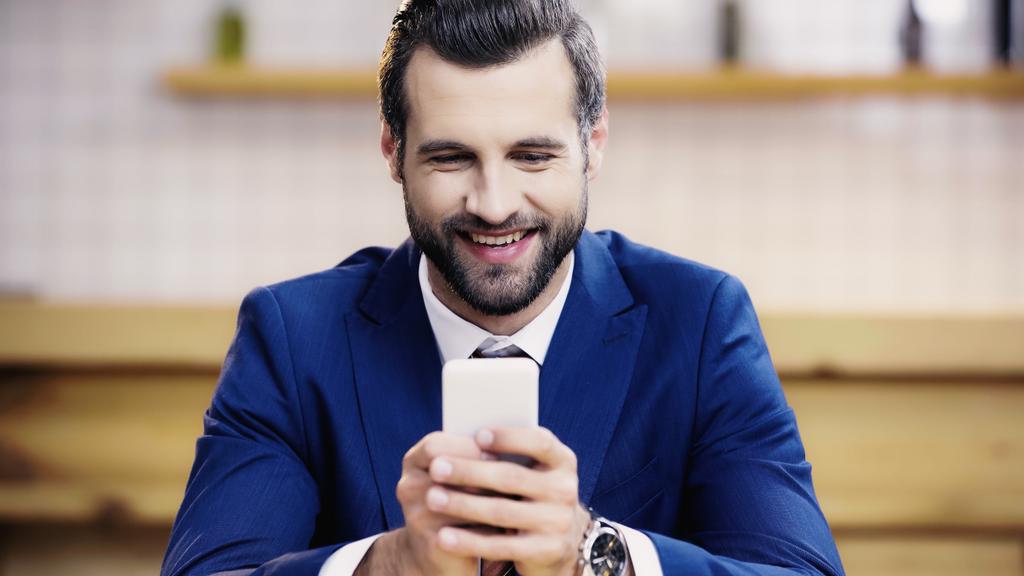 hombre de negocios barbudo en traje sonriendo mientras mensajea en el teléfono móvil en la cafetería  - Foto, imagen