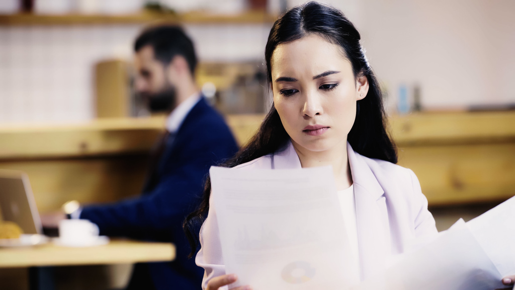focused asian businesswoman looking at documents near blurred businessman  - Photo, Image