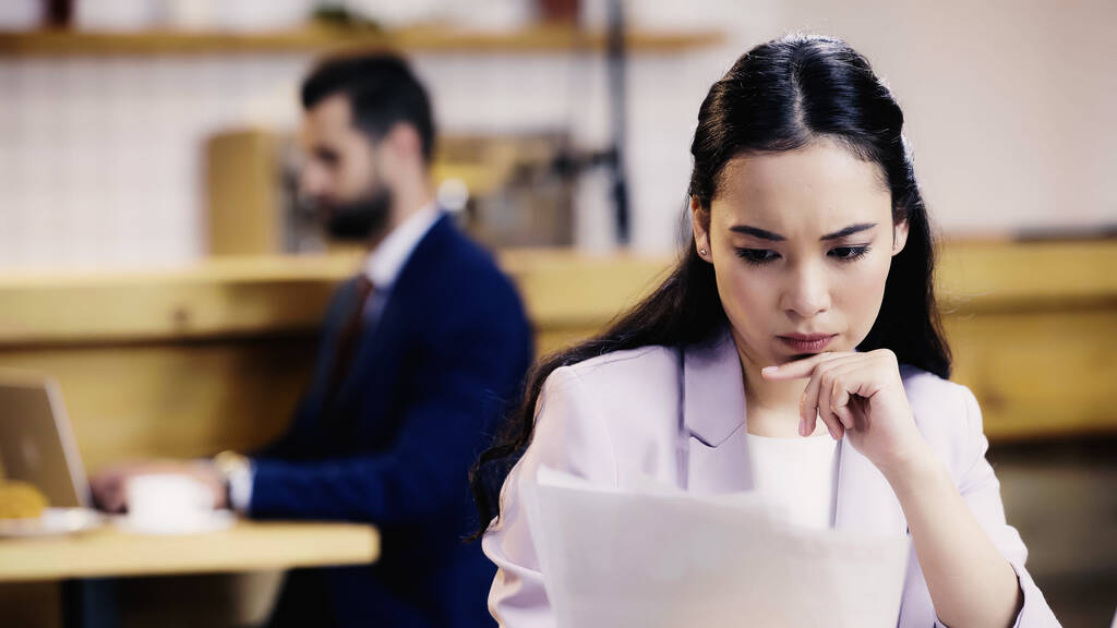 сфокусированная азиатская бизнесвумен смотрит на документы рядом с размытым бизнесменом на заднем плане в кафе  - Фото, изображение