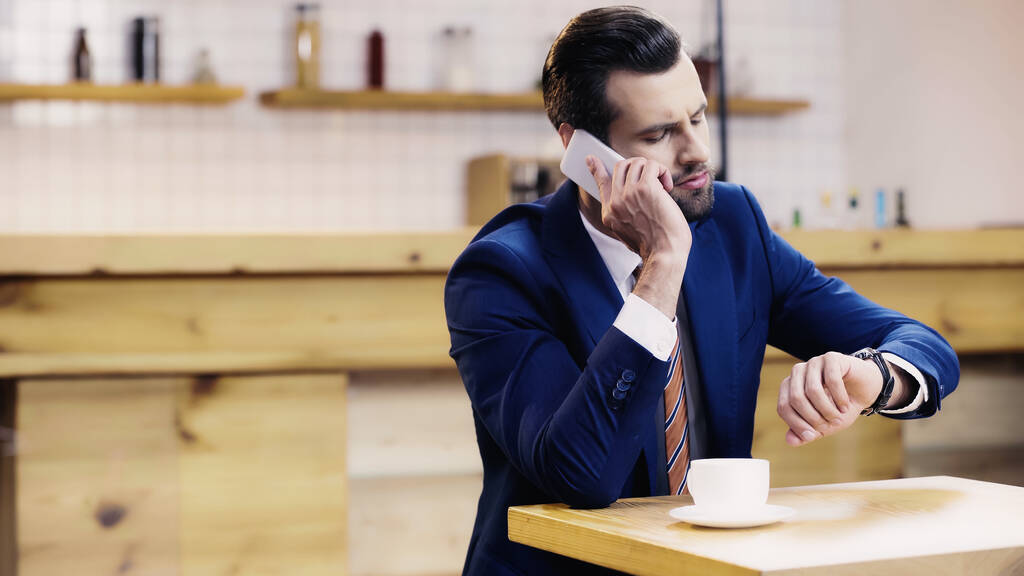 hombre de negocios barbudo en traje hablando en el teléfono inteligente y comprobando el tiempo en la cafetería - Foto, Imagen