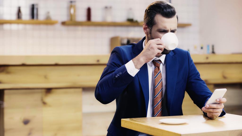 parrakas liikemies puvussa kahvin juominen ja älypuhelimen käyttö kahvilassa - Valokuva, kuva