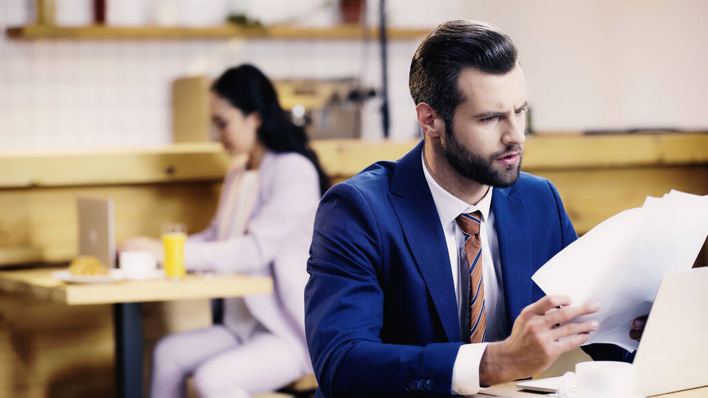 бородатий бізнесмен дивиться на документи біля ноутбука і розмита жінка в кафе
 - Фото, зображення