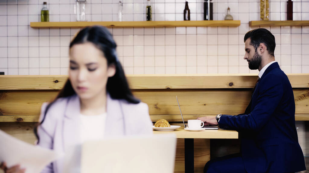 zakenman typen op laptop in de buurt wazig aziatische vrouw met documenten op de voorgrond  - Foto, afbeelding