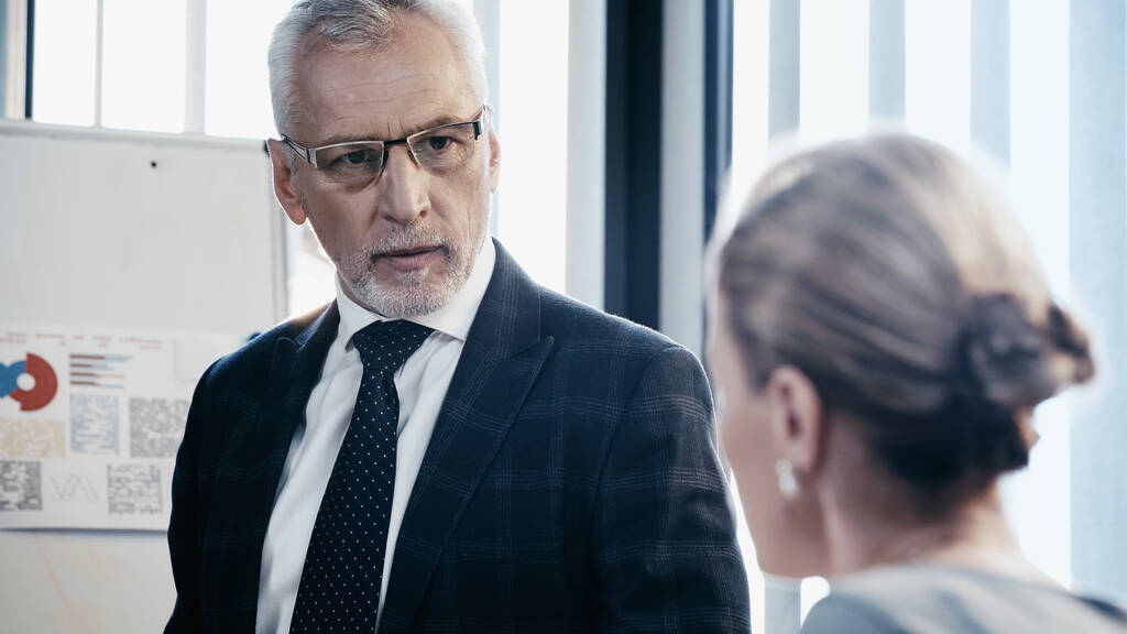 Uomo d'affari maturo in occhiali da vista guardando collega offuscata vicino flip chart in ufficio  - Foto, immagini