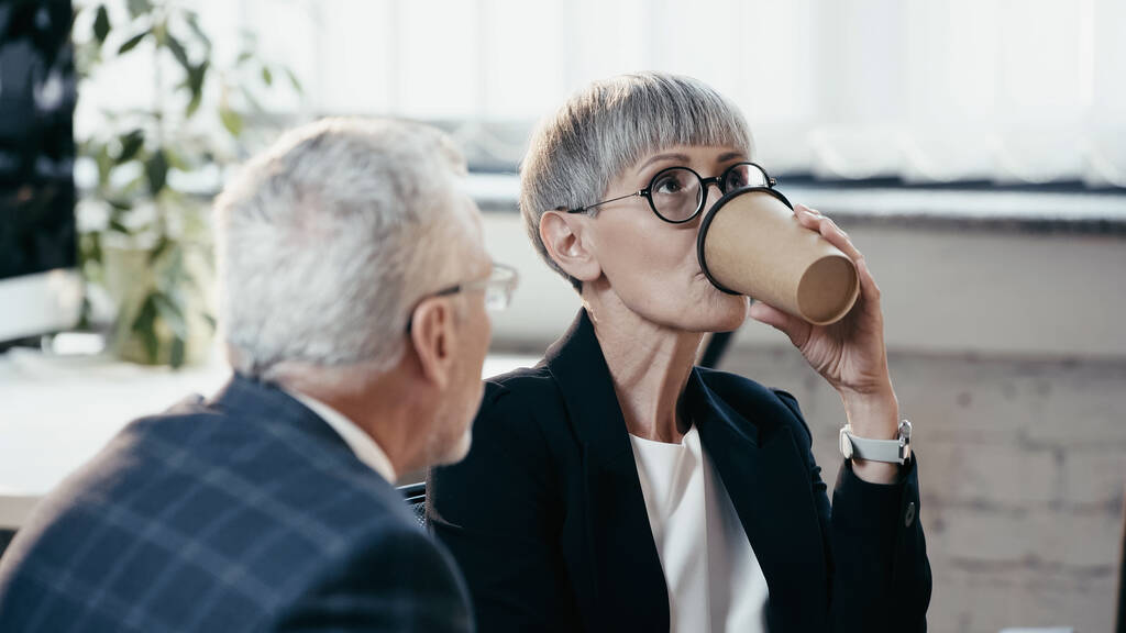 Mulher de negócios madura bebendo café para chegar perto confuso colega no escritório  - Foto, Imagem