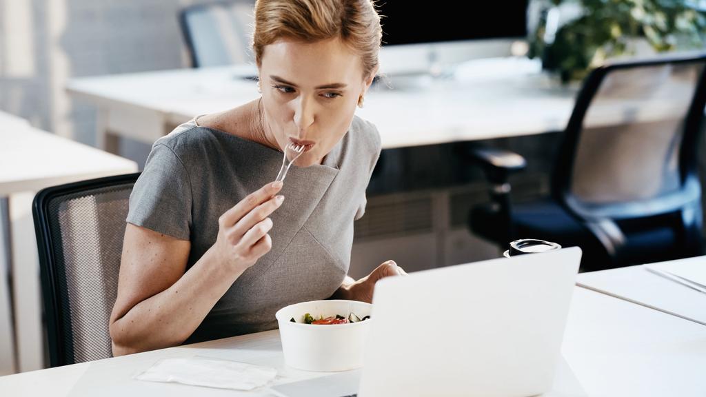 Donna d'affari mangiare insalata vicino al caffè per andare e laptop offuscata in ufficio  - Foto, immagini