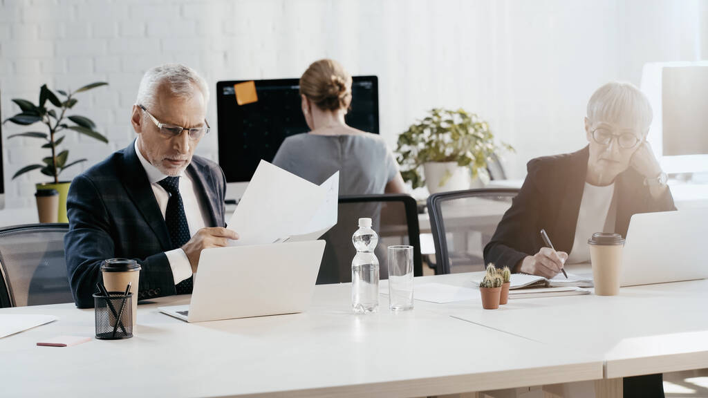 Зрелый бизнесмен с документами рядом с ноутбуком и коллегой, работающими в офисе  - Фото, изображение