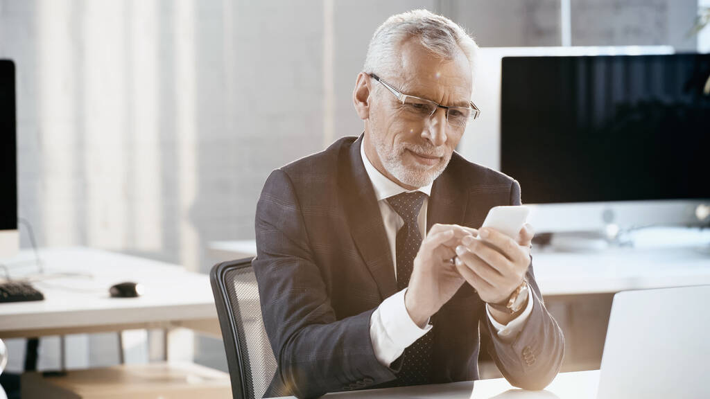 Усміхнений бізнесмен використовує мобільний телефон біля ноутбука в офісі
  - Фото, зображення