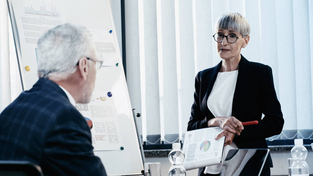 Middle aged businesswoman holding clipboard near colleague and flip chart in office  - Photo, Image